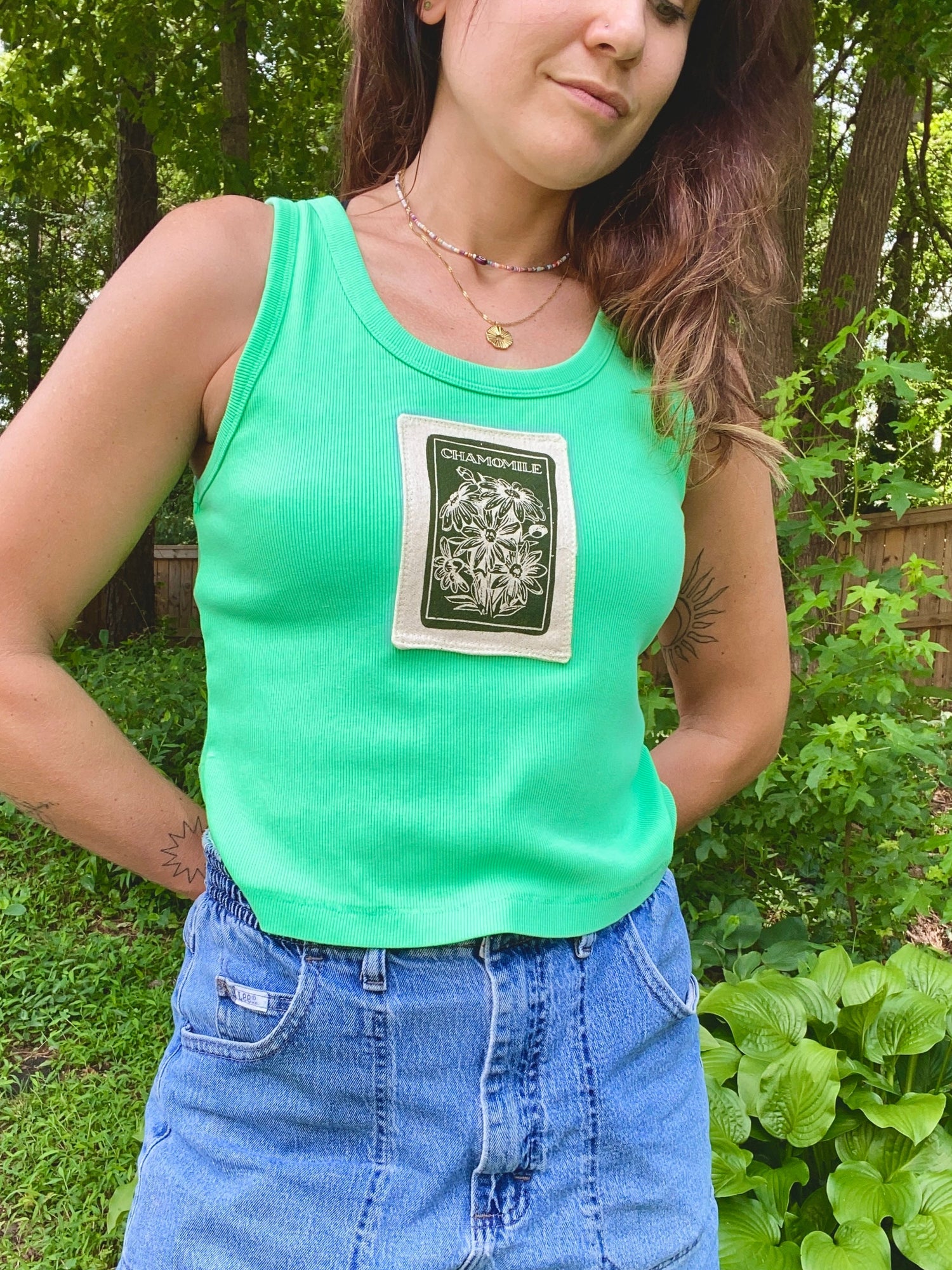 Person wearing a green tank top with a chamomile patch and blue shorts outdoors.